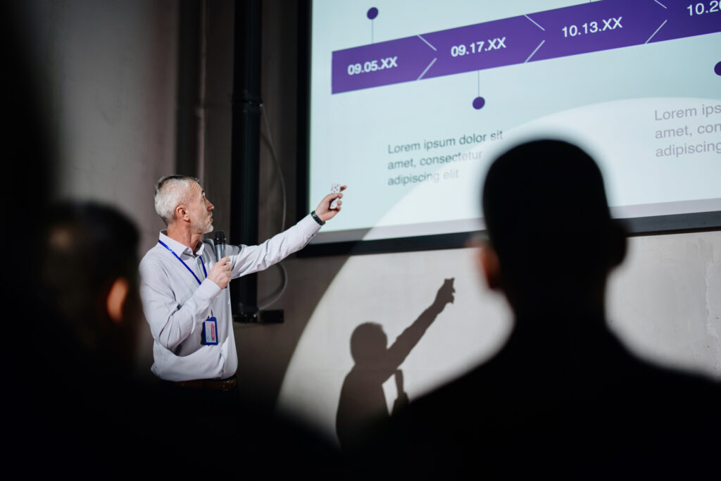 Successful male speaker presents on stage, uses remote control for presentation Showing Infographics
